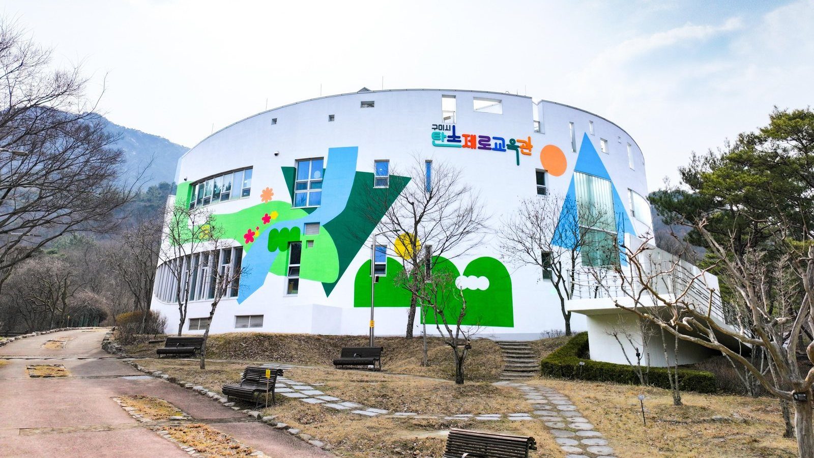산자락 아래 흰 원통형 건물. 벽에 초록·하늘색·노랑·주황 도형 벽화와 «구미시 탄소제로교육관» 글자가 있다