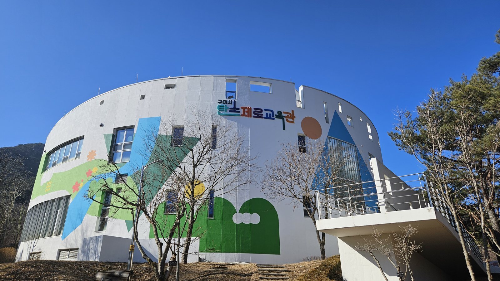 짙은 파란 하늘 아래 색색 도형 벽화가 그려진 흰 원통형 교육관 건물과 오른쪽 계단
